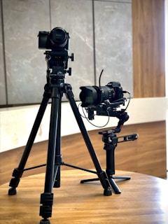 Two professional cameras mounted on tripods are placed on a wooden table. The background consists of a light-colored wall with wooden and stone textures.