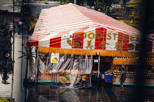 A street market tent with a red and white striped roof, labeled 'Food Street', featuring signs with images of food items and a transparent plastic covering on the sides. The market appears to be on a wet, reflective surface, possibly due to recent rain. People are walking with umbrellas, and one person is standing near the entrance. The atmosphere is lively with various items on display inside the tent.