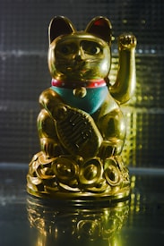 A gold figurine of a cat with one paw raised, often regarded as a symbol of luck and prosperity in many Asian cultures. It is adorned with a red collar and turquoise-colored bib, standing on a reflective surface with a textured gray background.