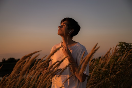 A person wearing earphones stands amidst tall grass under a warmly lit sky, casting a soft glow on their face. The scene conveys a calm and contemplative atmosphere as the person gazes upward.