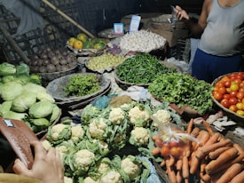 A bustling market stall displays an array of fresh produce including cabbages, beetroots, papayas, grapes, garlic, green chilies, cilantro, tomatoes, cauliflowers, and carrots. The produce is arranged in baskets and is vibrant in color, while a vendor is partially visible on the side. A hand holding a wallet suggests a transaction about to occur.