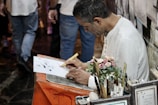 A person is sitting at a small table writing on a piece of paper with a calligraphy pen. The individual is focused on their work, surrounded by various writing tools and a small bouquet of artificial roses. In the background, there are framed pieces and people walking.