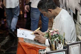 A person is sitting at a small table writing on a piece of paper with a calligraphy pen. The individual is focused on their work, surrounded by various writing tools and a small bouquet of artificial roses. In the background, there are framed pieces and people walking.