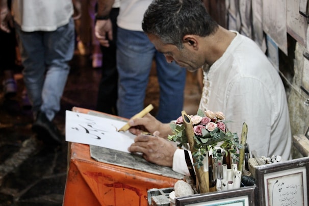 A person is sitting at a small table writing on a piece of paper with a calligraphy pen. The individual is focused on their work, surrounded by various writing tools and a small bouquet of artificial roses. In the background, there are framed pieces and people walking.