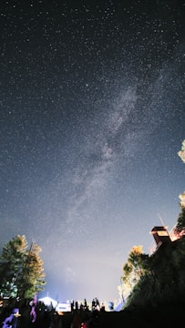 A starry night sky with the Milky Way visible in the background. Silhouettes of people and trees are in the foreground, with hints of light suggesting a lively gathering. A small structure or lookout tower is perched on the right side, illuminated by artificial lighting.