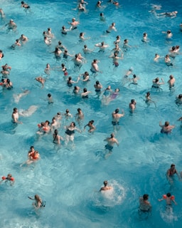 A crowded swimming pool with numerous people enjoying the water. Many individuals are swimming, splashing, and interacting with each other, wearing a variety of swimwear. The water is clear and has varying shades of blue.