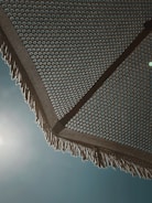 A woven beach umbrella is seen from underneath, with a detailed geometric pattern creating an intricate design. The sun is shining brightly behind it, casting subtle light and shadows. The umbrella's fringe is visible at the edges, with a clear blue sky as a backdrop.