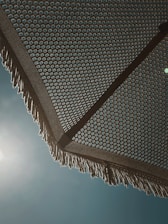 A woven beach umbrella is seen from underneath, with a detailed geometric pattern creating an intricate design. The sun is shining brightly behind it, casting subtle light and shadows. The umbrella's fringe is visible at the edges, with a clear blue sky as a backdrop.