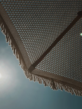 A woven beach umbrella is seen from underneath, with a detailed geometric pattern creating an intricate design. The sun is shining brightly behind it, casting subtle light and shadows. The umbrella's fringe is visible at the edges, with a clear blue sky as a backdrop.
