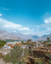 A scenic landscape featuring a large residential house surrounded by lush green trees, set against a backdrop of majestic mountains with snow-capped peaks under a clear blue sky. The foreground reveals a garden area with trees casting shadows on the ground.