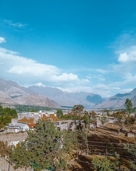A scenic landscape featuring a large residential house surrounded by lush green trees, set against a backdrop of majestic mountains with snow-capped peaks under a clear blue sky. The foreground reveals a garden area with trees casting shadows on the ground.