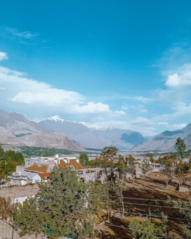 A scenic landscape featuring a large residential house surrounded by lush green trees, set against a backdrop of majestic mountains with snow-capped peaks under a clear blue sky. The foreground reveals a garden area with trees casting shadows on the ground.