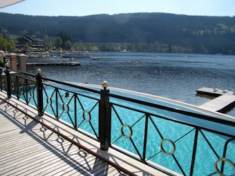 A serene lakeside view features a wooden deck with metal railings overlooking a swimming pool and a calm, expansive lake. In the distance, lush green hills and a collection of buildings can be seen. A few boats are scattered across the water, and a dock extends into the lake.