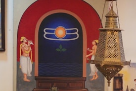 A decorative wall painting depicts two traditional Indian figures standing on either side of a large archway. The figures are wearing traditional attire and are performing rituals. In the center of the archway is a symbolic design featuring a sun and celestial motifs. A large, ornate brass lantern hangs from the ceiling in front of the painting.