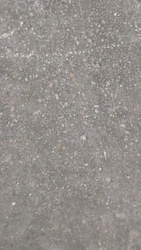 A concrete surface with a rough texture, featuring small pebbles and stones embedded within it. The surface has a gray color overall with specks of brown, beige, and white.