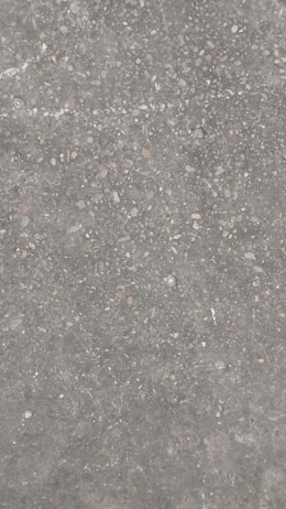 A concrete surface with a rough texture, featuring small pebbles and stones embedded within it. The surface has a gray color overall with specks of brown, beige, and white.