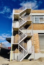 A metal fire escape with a zigzag pattern is attached to the side of a brick building. The building features large industrial windows, and the sky is bright blue with scattered clouds.