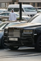 A luxurious black car with a distinctive front grille is parked in an urban environment. A man in traditional attire can be seen walking in the background, and several other vehicles, including a white van, are also visible.