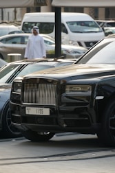 A luxurious black car with a distinctive front grille is parked in an urban environment. A man in traditional attire can be seen walking in the background, and several other vehicles, including a white van, are also visible.