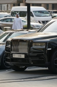 A luxurious black car with a distinctive front grille is parked in an urban environment. A man in traditional attire can be seen walking in the background, and several other vehicles, including a white van, are also visible.