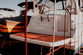 The image depicts the back of a white boat with the name written in Cyrillic script, moored alongside other boats. There is a nautical atmosphere with visible boat decks and some sunshine casting shadows.