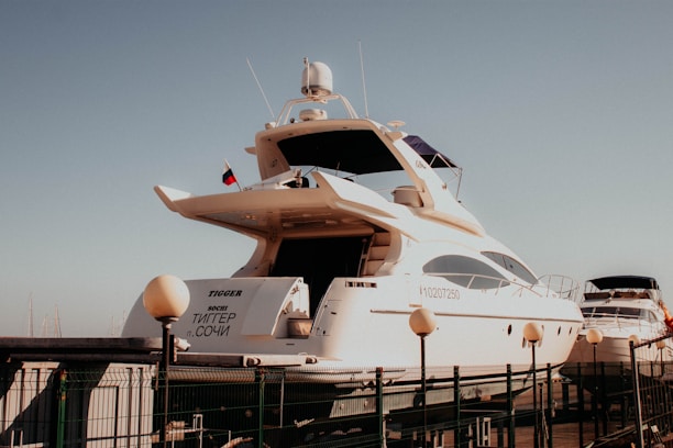 A large, white yacht is docked at a marina, with several smaller boats visible in the background. The yacht has a sleek design with multiple decks and a visible name on the back. A flag is flying atop, and a fence surrounds the dock area.
