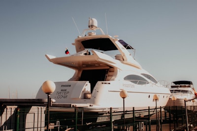 A large, white yacht is docked at a marina, with several smaller boats visible in the background. The yacht has a sleek design with multiple decks and a visible name on the back. A flag is flying atop, and a fence surrounds the dock area.