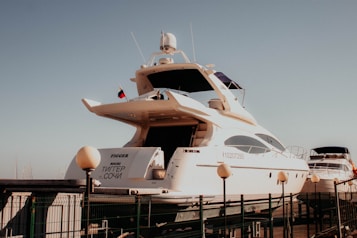 A large, white yacht is docked at a marina, with several smaller boats visible in the background. The yacht has a sleek design with multiple decks and a visible name on the back. A flag is flying atop, and a fence surrounds the dock area.