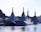 A group of large, modern naval ships is docked at a harbor. The vessels have angular, stealthy designs with tall masts and various equipment. Surrounding buildings and greenery are visible in the background, under a clear sky.
