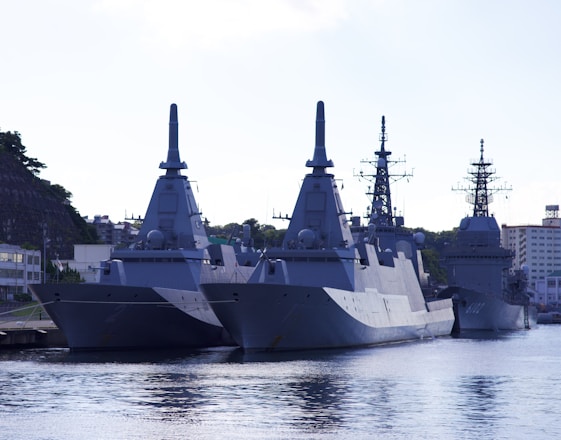 A group of large, modern naval ships is docked at a harbor. The vessels have angular, stealthy designs with tall masts and various equipment. Surrounding buildings and greenery are visible in the background, under a clear sky.