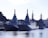 A group of large, modern naval ships is docked at a harbor. The vessels have angular, stealthy designs with tall masts and various equipment. Surrounding buildings and greenery are visible in the background, under a clear sky.