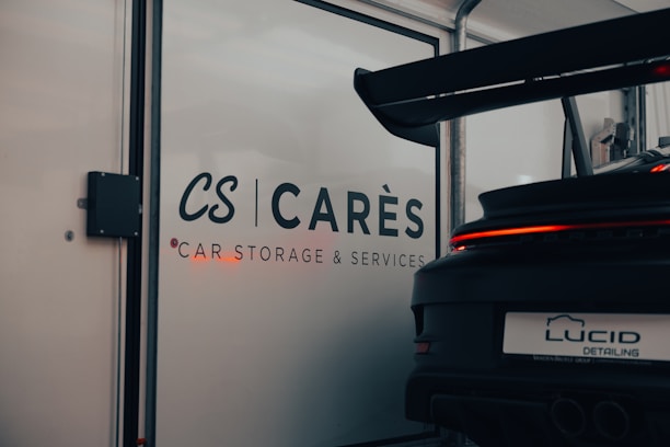 A garage with signage displaying the words 'CARÈS Car Storage & Services' on a glass door. Part of a sports car with a large rear spoiler, visible from the back, occupies the right side of the image, with 'LUCID Detailing' on its license plate.