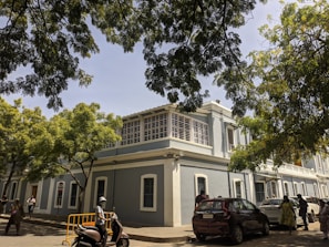 A colonial-style building with light blue walls, featuring large windows and a rooftop balcony. The scene is shaded by large trees, and there are several people casually walking and a person riding a scooter. Cars are parked along the street, and a traffic sign is visible.