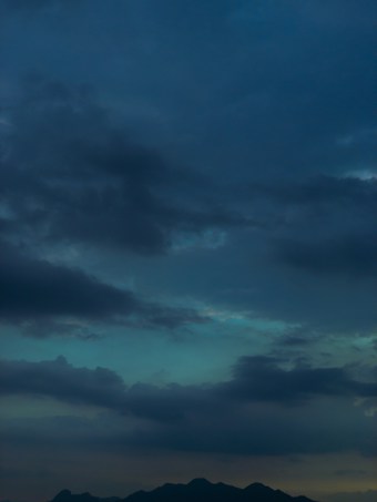 A landscape with a dark, cloudy sky transitioning from twilight to night, with hints of blue and dark gray. Silhouetted mountains create a subtle contrast at the bottom of the image.