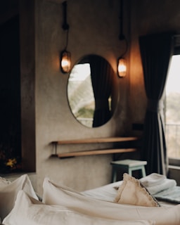 A cozy bedroom featuring soft pillows on a neatly made bed with a textured beige throw blanket. The room includes a round mirror mounted on a concrete wall, flanked by two warm-toned wall sconces. A small, wooden shelf and a stool are positioned beneath the mirror. Dark curtains partially drawn open reveal a hint of natural light entering through a window.