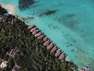 An aerial view of a tropical beach resort featuring overwater bungalows arranged in a line along the coastline. The turquoise water is clear, with visible coral patches. Lush green vegetation and palm trees cover the land area, providing a contrast to the sandy beach and blue sea.