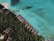 An aerial view of a tropical beach resort featuring overwater bungalows arranged in a line along the coastline. The turquoise water is clear, with visible coral patches. Lush green vegetation and palm trees cover the land area, providing a contrast to the sandy beach and blue sea.