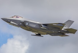 A modern military jet flies against a backdrop of cloudy sky. The aircraft is sleek with a gray camouflaged design, showcasing aerodynamic features and weapon mounts.