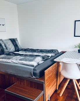 A minimalist bedroom with a wooden platform bed and patterned bedding, adjacent to a simple white desk and modern chair. On the wall, two framed artworks are visible, and a small green plant sits on the desk.