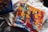 A vibrant and abstract painting on canvas featuring a mix of bright colors including red, yellow, blue, and white. The painting includes bold brush strokes and geometric shapes with a textured surface, creating a dynamic composition. Another canvas with similar abstract artwork is partially visible nearby, all placed on a soft, light-colored fabric background.
