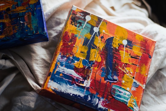 A vibrant and abstract painting on canvas featuring a mix of bright colors including red, yellow, blue, and white. The painting includes bold brush strokes and geometric shapes with a textured surface, creating a dynamic composition. Another canvas with similar abstract artwork is partially visible nearby, all placed on a soft, light-colored fabric background.