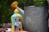 A bottle of yellow mustard sits on a surface with blue paint dripping down its sides. It is placed near a large, weathered metal watering can with some handwritten numbers on it. In the background, there is greenery visible, suggesting an outdoor setting.