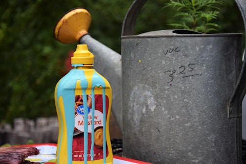 A bottle of yellow mustard sits on a surface with blue paint dripping down its sides. It is placed near a large, weathered metal watering can with some handwritten numbers on it. In the background, there is greenery visible, suggesting an outdoor setting.