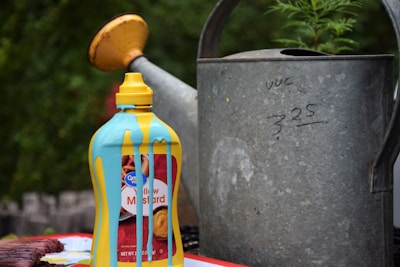 A bottle of yellow mustard sits on a surface with blue paint dripping down its sides. It is placed near a large, weathered metal watering can with some handwritten numbers on it. In the background, there is greenery visible, suggesting an outdoor setting.