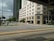 A hospital building with multiple floors and large windows located at a street corner. The scene includes a wide road with yellow and white lane markings, a pedestrian walking on a sidewalk surrounded by greenery, and a partly cloudy sky. Another tall building is visible in the background.
