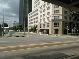 A hospital building with multiple floors and large windows located at a street corner. The scene includes a wide road with yellow and white lane markings, a pedestrian walking on a sidewalk surrounded by greenery, and a partly cloudy sky. Another tall building is visible in the background.