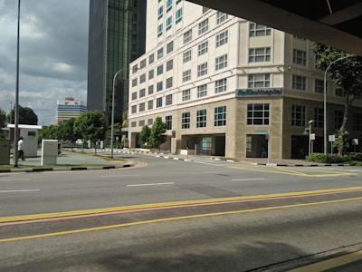 A hospital building with multiple floors and large windows located at a street corner. The scene includes a wide road with yellow and white lane markings, a pedestrian walking on a sidewalk surrounded by greenery, and a partly cloudy sky. Another tall building is visible in the background.