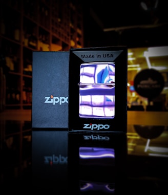 A Zippo lighter with a purple flame is displayed in the foreground. The lighter is encased in a black box with the text 'Made in USA' at the top. The black box and its reflection are clearly visible on a surface, likely glass or polished wood. The background is slightly blurred, showing shelves and a blurred circular sign with indistinct text, suggesting a store or display environment.