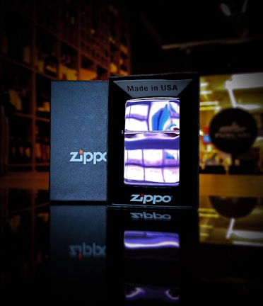 A Zippo lighter with a purple flame is displayed in the foreground. The lighter is encased in a black box with the text 'Made in USA' at the top. The black box and its reflection are clearly visible on a surface, likely glass or polished wood. The background is slightly blurred, showing shelves and a blurred circular sign with indistinct text, suggesting a store or display environment.