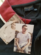 A clothing tag with the brand name 'Kleinigkeit' is attached to a garment featuring a red and black color scheme. The tags display images of a man, one sitting outdoors wearing sunglasses and a white t-shirt, while the other shows him indoors with a hand to his head.